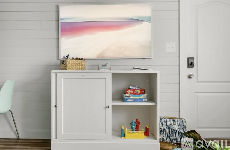 A white cabinet with a painting above it and a white chair to the left.