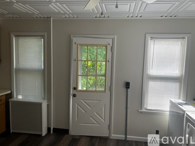 A room with a white door, two windows with blinds, and a radiator on the left.