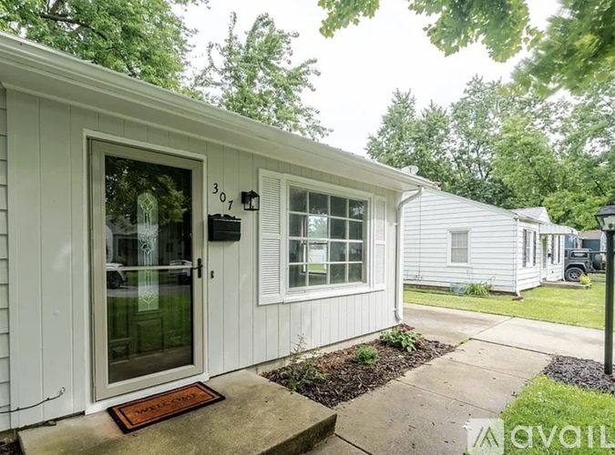 A small white house with a glass door and a window is surrounded by greenery.