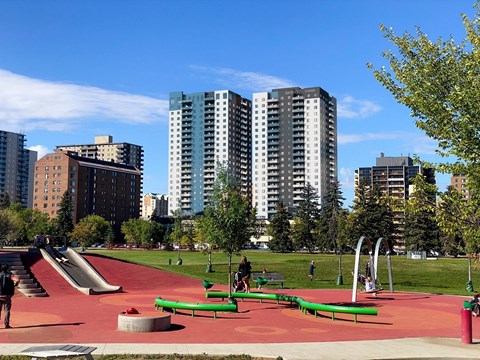 A playground with a red surface and green benches is surrounded by tall buildings.