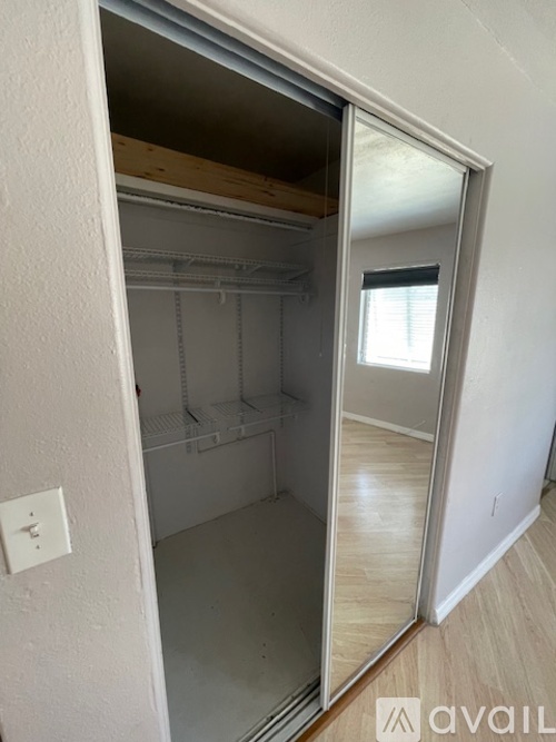 A white door with a mirror on the side is open to a room with a wooden floor and a window.