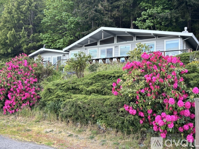 A house with a grey roof and white walls is surrounded by greenery and pink flowers.