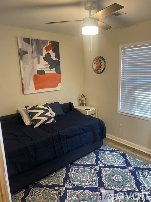 A bedroom with a bed, a ceiling fan, and a painting on the wall.