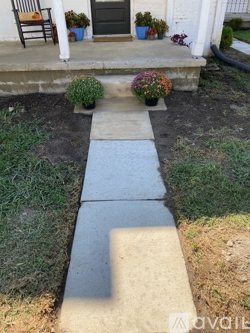 A concrete walkway leads to a house with a chair and potted plants on the porch.