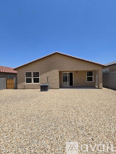A house with a brown roof and tan walls is surrounded by a gravel lot.