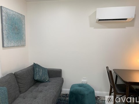 A living room with a grey couch, a blue ottoman, a painting on the wall, and a table with a lamp.