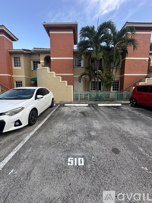 A white car is parked in front of a red car and a building with a palm tree.
