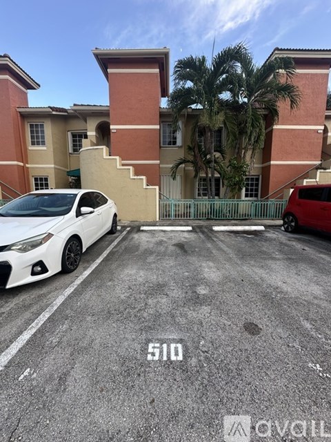 A white car is parked in front of a red car and a building with a palm tree.