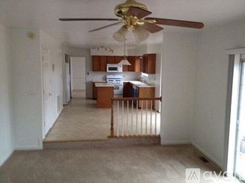 A living room with a ceiling fan and a kitchen area in the background.