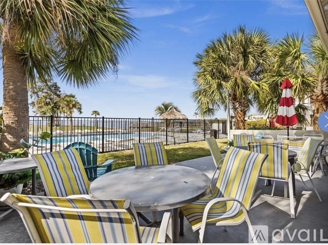 A patio with striped chairs and a table is set up with a pool and palm trees in the background.