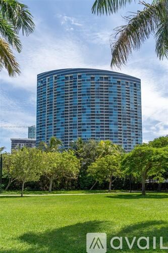 A tall building with a blue tinted glass facade is surrounded by trees and grass.