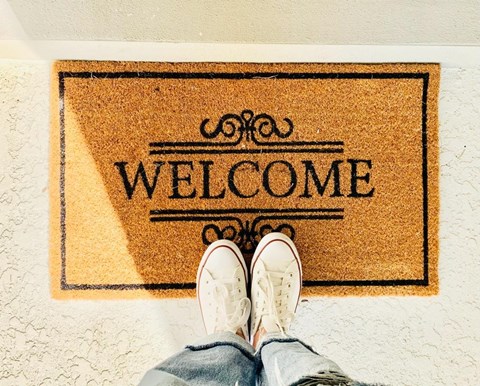A pair of white sneakers is stepping on a doormat that says "Welcome".