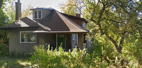 A small house with a brown roof and a chimney is surrounded by greenery.