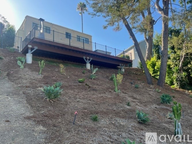 A house with a balcony is built on a slope with some plants and trees around it.