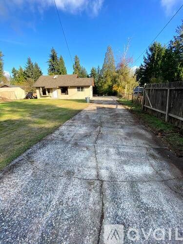 A concrete driveway leads to a house with a fence on one side and trees on the other.