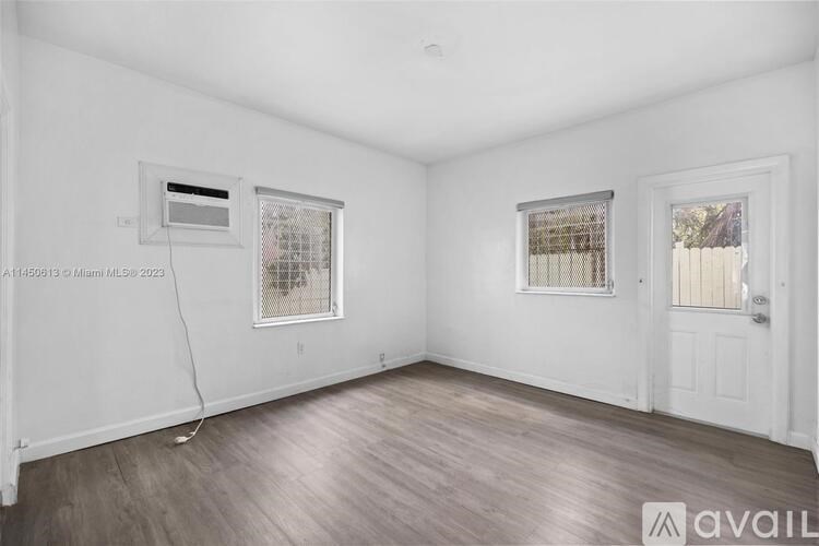 A room with white walls and wooden flooring, featuring a window with a view of a fence and a door on the right.