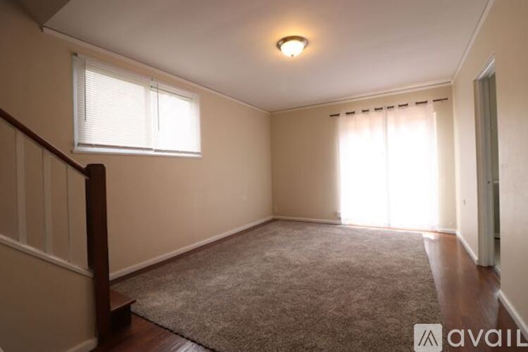 A room with a carpeted floor, a window with blinds, and a wooden staircase.