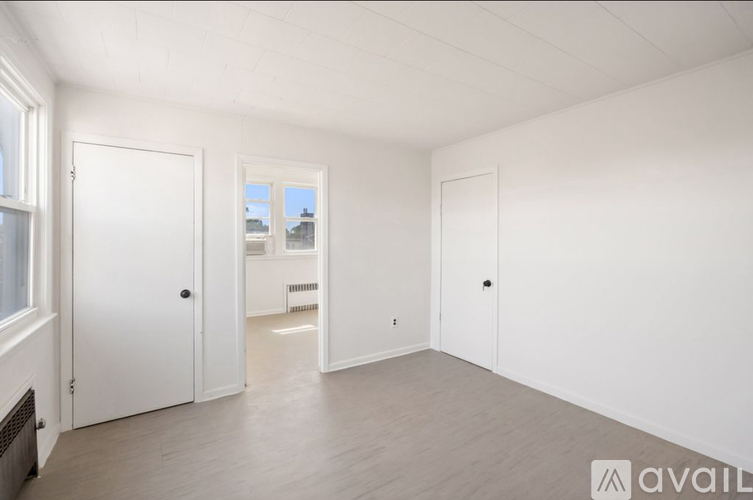A spacious room with white walls and wooden flooring, featuring a fireplace, a window, and a door leading to another room.