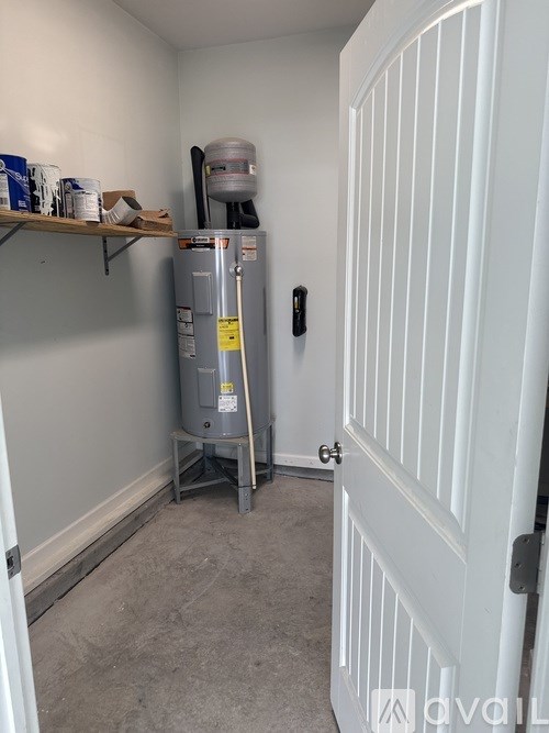 A room with a white door and a grey floor with a water heater and a shelf with paint cans.