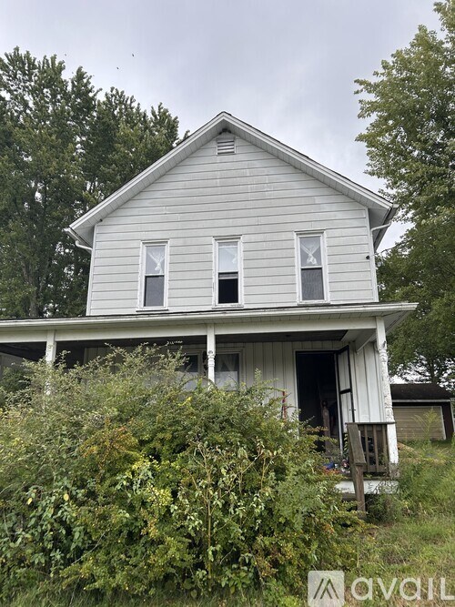 A white house with a boarded up window sits in front of a green bush.