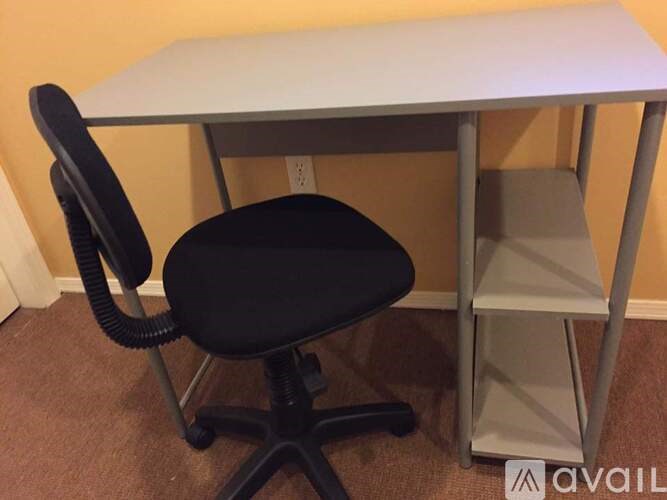 A black chair with a black seat cushion is in front of a white desk with a grey shelf.
