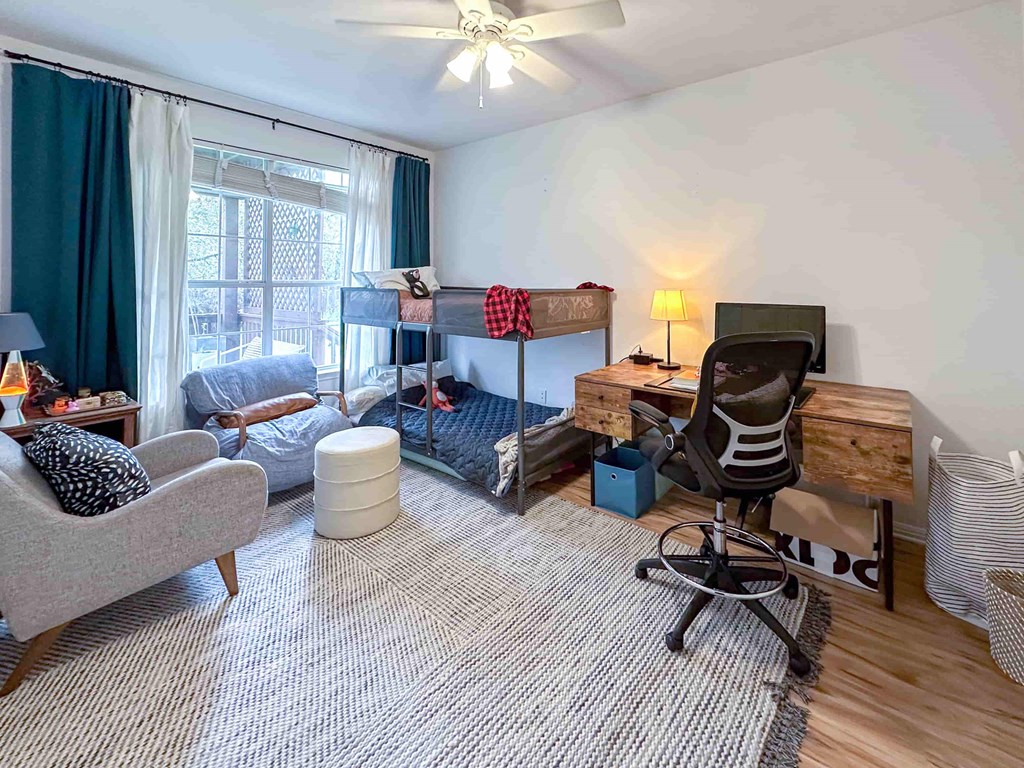 A room with a grey sofa, a bed with a red and black blanket, a desk with a chair, and a window with curtains.