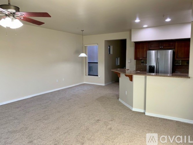 A spacious living room with a ceiling fan and a kitchen area with a refrigerator.