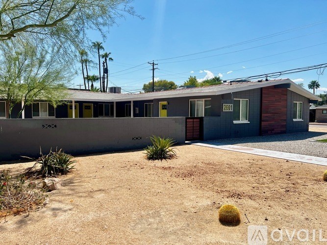 A house with a grey and red exterior is surrounded by a gravel driveway and desert vegetation.