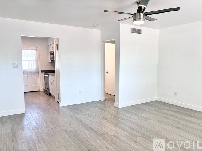 A spacious room with a ceiling fan and light fixture, and a kitchen visible through the doorway.