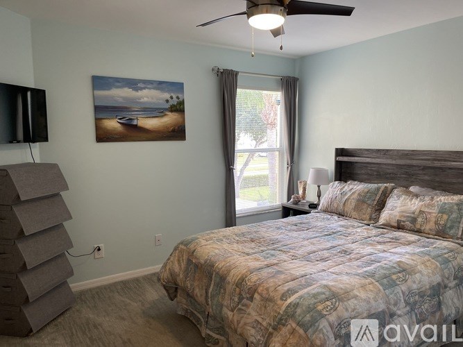 A bedroom with a bed, a ceiling fan, and a painting of a beach scene on the wall.