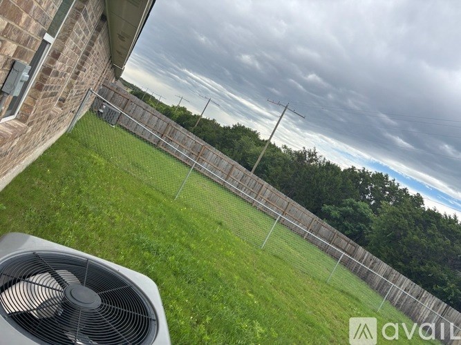 A fan is in the foreground of a grassy area with a fence and trees in the background.