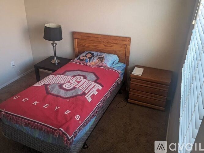 A bed with a red Ohio State blanket is in a room with a lamp and a nightstand.