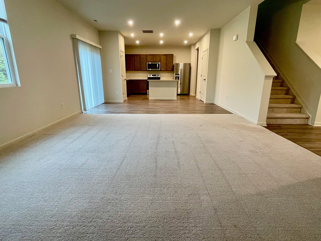 A spacious living room with a carpeted floor and a staircase on the right side.