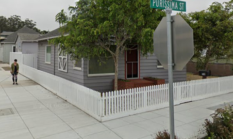 A person walks down a sidewalk past a house with a white picket fence.