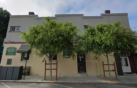 A building with a green sign on the left side and a tree in front of it.