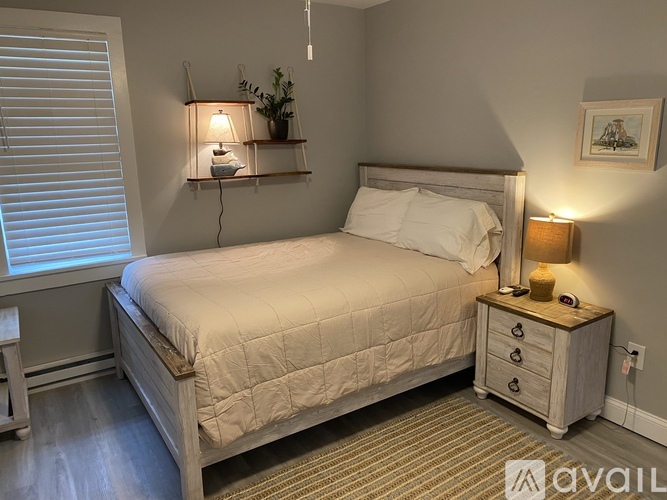 A well-lit bedroom with a neatly made bed, a nightstand with a lamp, and a small table beside the window.