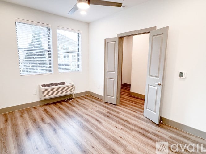 A room with a wooden floor, a window with blinds, a white air conditioner, and two doors.