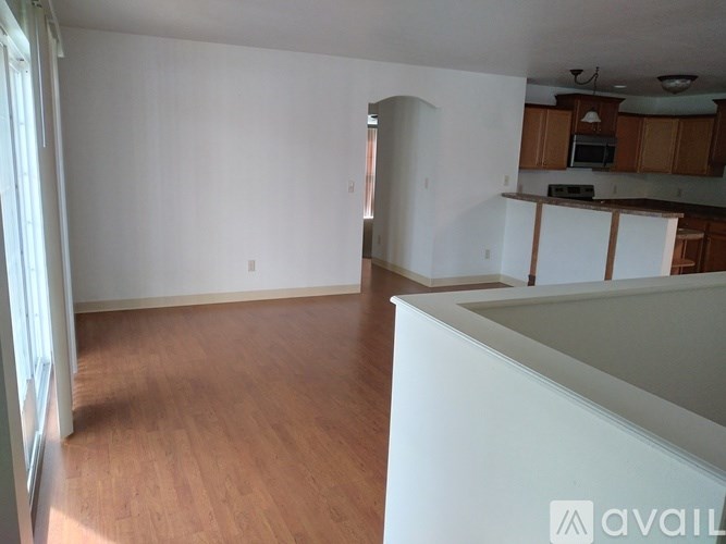 A room with wooden floors and white walls, with a kitchen area visible in the background.
