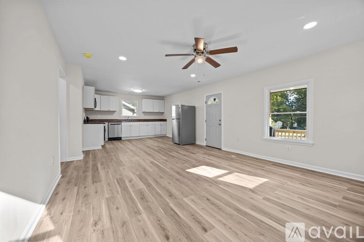 A spacious room with a ceiling fan and lighting, hardwood flooring, and a window with a view of trees.