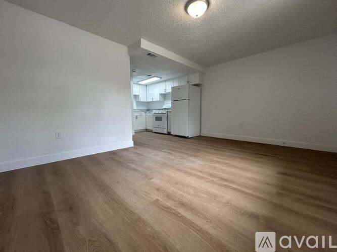 A room with wooden flooring and a white wall with a fridge in the background.