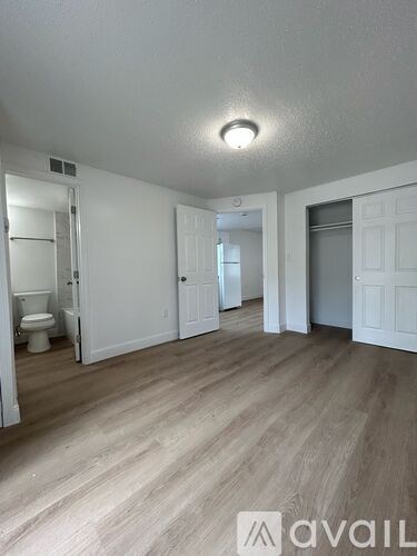 A room with white walls and wooden flooring, with a toilet visible in the background.