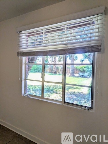 A window with white blinds and a view of a green lawn and trees outside.