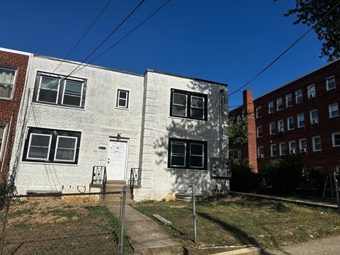 A white two-story building with a grey roof and a white door is surrounded by a grassy area and a sidewalk.