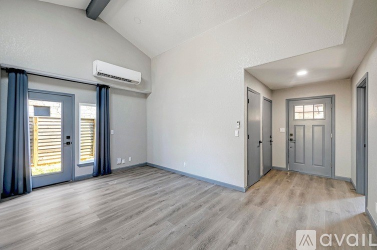 A spacious room with wooden flooring and two doors, one of which is open to a balcony with blinds.