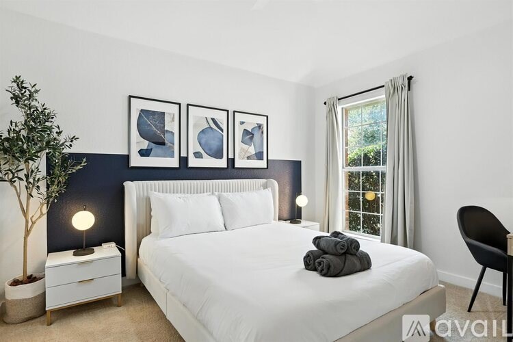 A bedroom with a white bed, a white nightstand with a lamp, a plant, and three framed pictures on the wall.