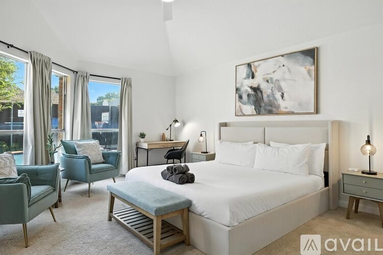 A well-lit bedroom with a large bed, a chair, a nightstand, and a painting on the wall.