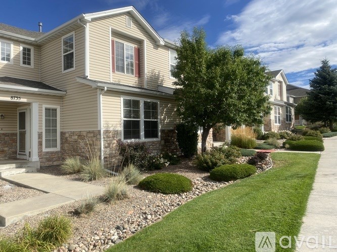 A beige house with a white door and windows is surrounded by a well-manicured lawn and landscaping.
