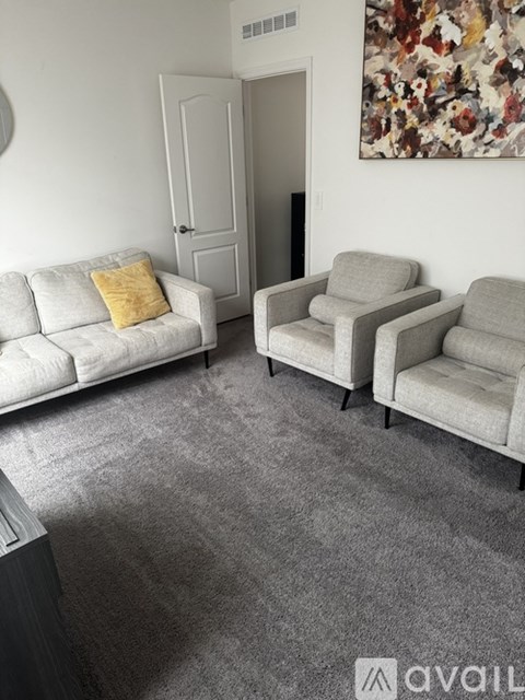 A living room with a grey carpet, a white sofa, a yellow pillow, a grey chair and a painting on the wall.
