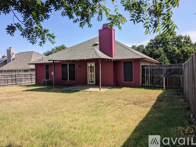 A red house with a brown roof and a chimney is surrounded by a fence and has a grassy yard.