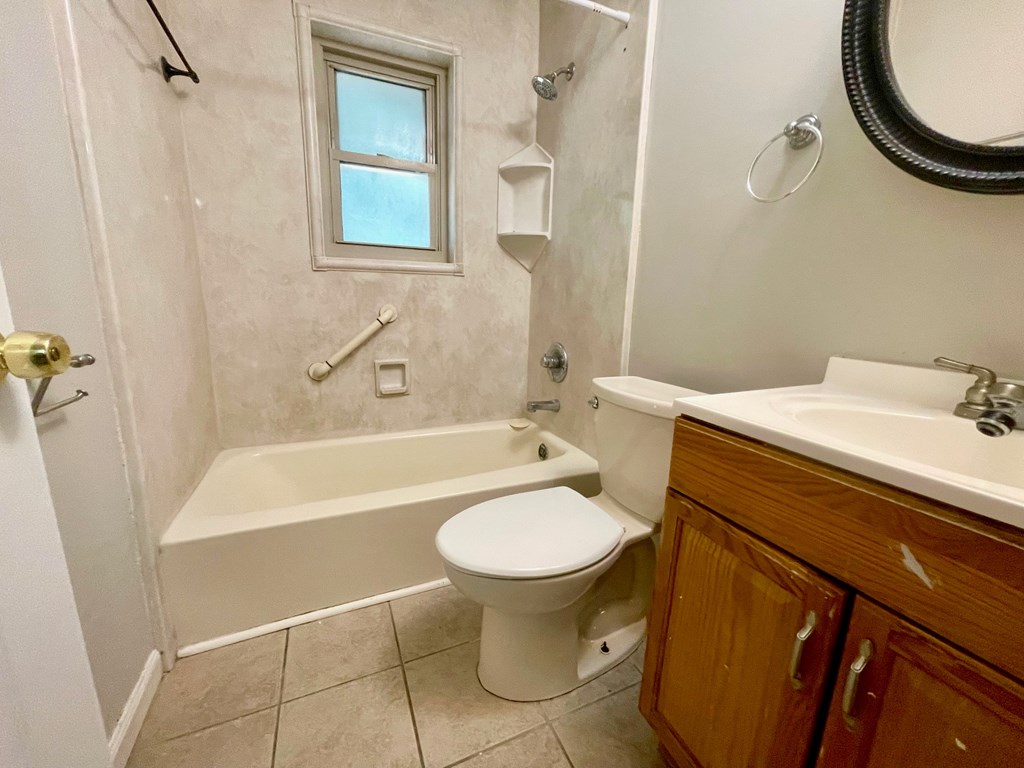 A white toilet sits in a bathroom with a white tub and a window in the shower.